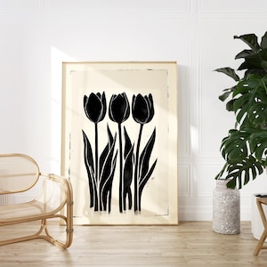 May include: A framed art print featuring a black and white illustration of three tulips. The tulips have long stems and leaves, set against a cream-colored background. The artwork is displayed on a wooden floor, with a rattan chair and a potted plant in the foreground.