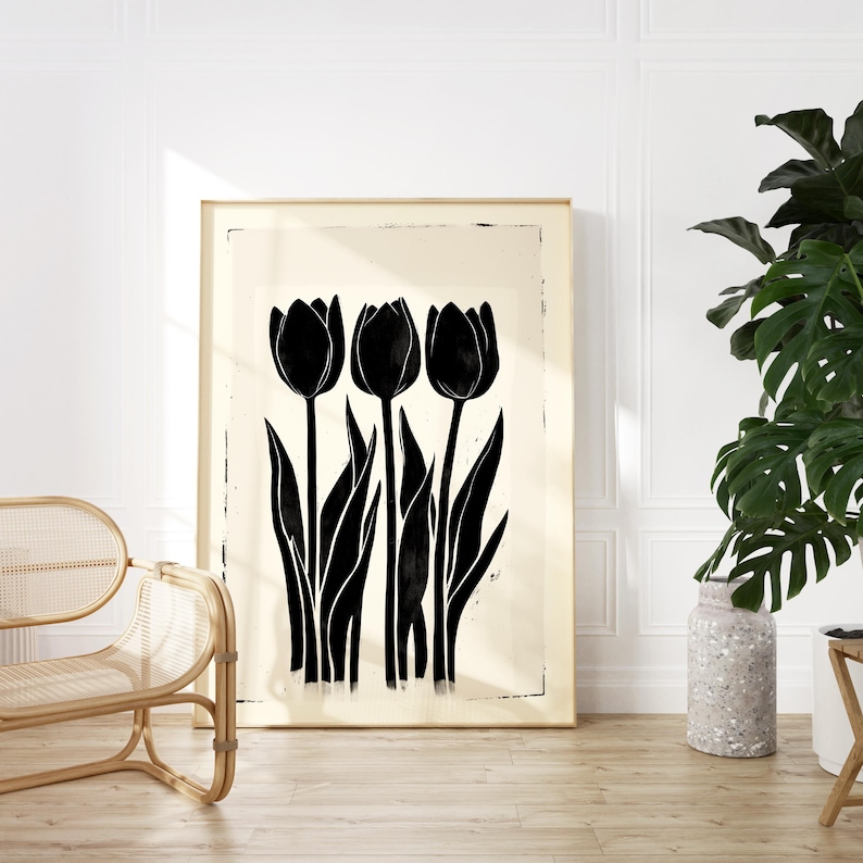 May include: A framed art print featuring a black and white illustration of three tulips. The tulips have long stems and leaves, set against a cream-colored background. The artwork is displayed on a wooden floor, with a rattan chair and a potted plant in the foreground.