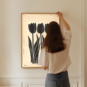 May include: A framed art print featuring a black and white tulip design. The artwork showcases three stylized tulips with long, slender leaves against a cream-colored background. The frame is a light wood color, adding a natural touch to the floral art.