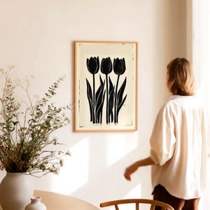 May include: A framed art print featuring a black and white illustration of three tulips. The tulips have long stems and leaves, set against a cream-colored background. The artwork is displayed in a light brown wooden frame, adding a touch of natural warmth to the floral design.