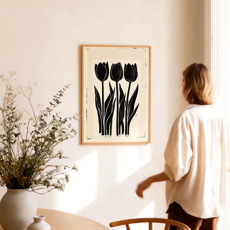 May include: A framed art print featuring a black and white illustration of three tulips. The tulips have long stems and leaves, set against a cream-colored background. The artwork is displayed in a light brown wooden frame, adding a touch of natural warmth to the floral design.