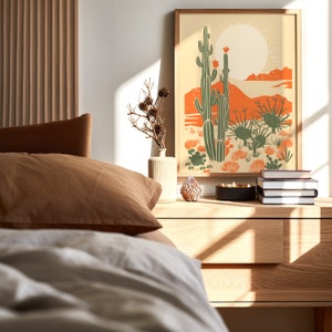 May include: A bedroom interior with a wooden nightstand, a white and brown bed with a brown pillow, a vase with dried branches, a stack of books, a candle, and a framed print of a desert landscape with cacti and a sun.