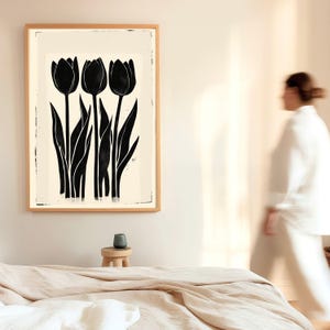 May include: A framed art print featuring three black tulip silhouettes against a cream background. The artwork is in a natural wood frame, creating a minimalist and modern aesthetic. The image is in a bedroom setting.