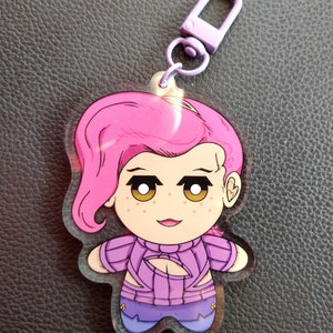 May include: A pink and purple anime character keychain with a purple sweater and blue pants. The keychain has a purple metal clasp.