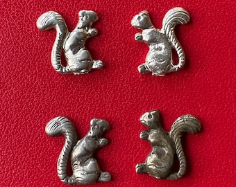 May include: Four small, silver-colored squirrel figurines arranged on a bright red background. Each squirrel is depicted in a standing pose with a bushy tail. The figurines appear to be made of metal.