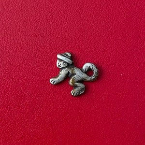 Könnte beinhalten: Kleine, silberfarbene Affenfigur mit Hut. Der Affe ist in einer Laufpose mit eingerolltem Schwanz dargestellt. Die Figur hat goldfarbene Akzente und ist vor einem roten Hintergrund platziert.