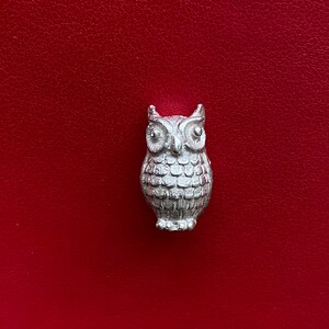 May include: A silver-colored owl figurine with detailed feather and eye features. The owl is centered against a red background. The figurine is a small decorative item.