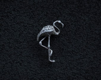 May include: A silver flamingo figurine, standing on one leg, with detailed feather textures. The bird's long neck curves gracefully, and its beak is slightly open. The figurine is set against a dark, textured background.