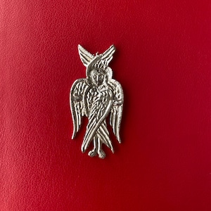 Może przedstawiać: Srebrny wisiorek w kształcie anioła z rozłożonymi skrzydłami i szczegółowymi piórami. Wisiorek jest umieszczony na czerwonym tle, podkreślając jego skomplikowany design i religijny motyw. Twarz anioła jest widoczna.