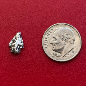 Puede incluir: Una pequeña punta de flecha plateada con una superficie texturizada, junto a una moneda de diez centavos estadounidense. La punta de flecha es triangular con una punta. La moneda de diez centavos presenta un perfil de un hombre y las palabras "Liberty" e "In God We Trust".