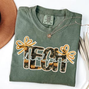 May include: Sage green Comfort Colors t-shirt with the words "TECH dialysis" in a decorative font. The letters are filled with a pattern of sunflowers and pumpkins. Gold bows accent the top of the words. A silver necklace with a pendant is on the shirt.