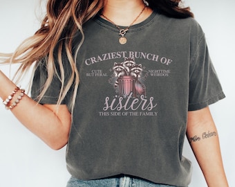 Puede incluir: Camiseta gris oscuro con el texto "CRAZIEST BUNCH OF sisters" y un gráfico de mapaches en un cubo de basura. La camiseta también tiene las frases "CUTE BUT FERAL" y "NIGHTTIME WEIRDOS".