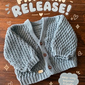 May include: A light blue long-sleeve cardigan with a textured bobble stitch pattern, a V-neckline, and three brown buttons. The text "PATTERN RELEASE!" and "easy • cozy beginner friendly" are visible, along with a small tag labeled "Handmade".
