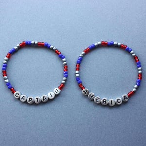 Puede incluir: Dos pulseras de cuentas con cuentas rojas, blancas y azules. Una pulsera dice "CAPTAIN" y la otra dice "AMERICA".