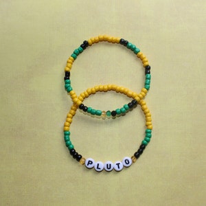 May include: Two beaded bracelets with black, yellow, and green beads. The smaller bracelet has the word "PLUTO" spelled out in white beads.