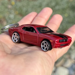 Può includere: Un'auto modello pressofusa rossa con strisce nere sul cofano, attaccata a un portachiavi argentato. L'auto ha ruote argentate dettagliate e una finitura lucida. L'auto misura circa 7.6 cm di lunghezza.