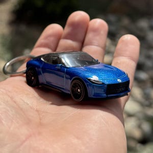 May include: A miniature blue sports car keyring with a silver key ring. The car has black wheels and a black roof. The car is held in a hand against a blurred background.
