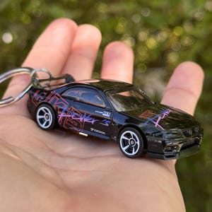 May include: Black die-cast model car keychain with white wheels and a rear spoiler. The car features pink and purple graphics and the text "Fighter". The keychain is held in a hand.