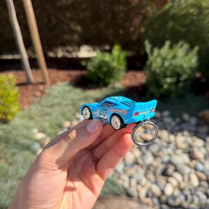 Może przedstawiać: Jasnoniebieski brelok do kluczy w kształcie zabawkowego samochodu z numerem 95 i napisem "Dinoco" z boku. Samoch&oacute;d ma białe koła i srebrny brelok. Tło przedstawia zieleń i słoneczne otoczenie na zewnątrz.
