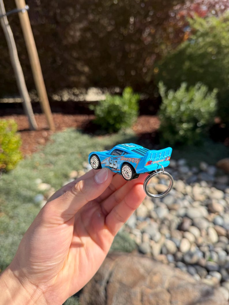 Może przedstawiać: Jasnoniebieski brelok do kluczy w kształcie zabawkowego samochodu z numerem 95 i napisem "Dinoco" z boku. Samoch&oacute;d ma białe koła i srebrny brelok. Tło przedstawia zieleń i słoneczne otoczenie na zewnątrz.