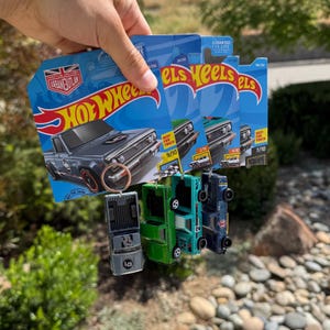 May include: A collection of Hot Wheels toy cars, including a gray pickup truck, held in front of their blue packaging. The packaging features the Hot Wheels logo and the text "URBAN OUTLAW". Several other toy cars in green and blue are also visible.