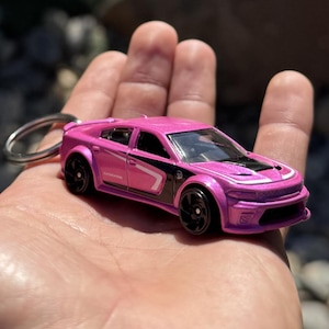 以下が含まれることがあります： ピンク色のダイキャスト製モデルカーキーホルダー。車は黒いホイール、ボンネットの黒と白のストライプ、側面の白いストライプが特徴です。「DODGE」の文字が車の側面に表示されています。