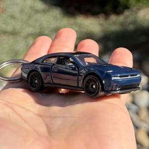 以下が含まれることがあります： ネイビーブルーのダイキャスト製モデルカーキーホルダー。 ミニカーは黒いホイールと黒い屋根が特徴です。 キーホルダーは、ぼやけた緑色の背景に対して手で持たれています。