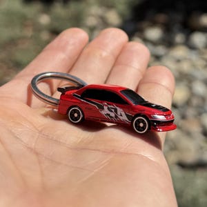 May include: A red toy car keyring with black detailing and a silver ring. The car features a detailed design with black and white graphics on the side and black wheels. A small spoiler is visible at the rear.