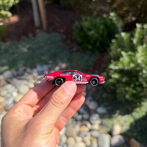May include: A small, red toy car with black wheels and the number 34 on the side. The car has a keychain attached and is held in a person's hand. The background is blurred with greenery and rocks.