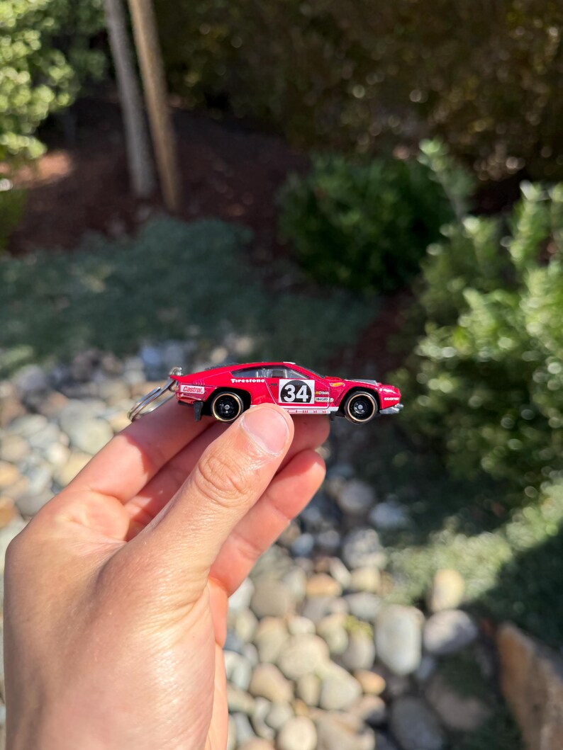 May include: A small, red toy car with black wheels and the number 34 on the side. The car has a keychain attached and is held in a person's hand. The background is blurred with greenery and rocks.