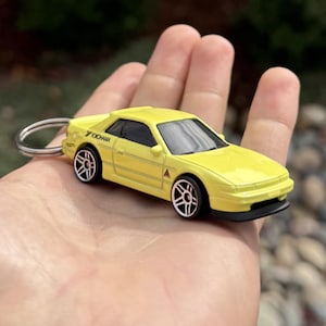 May include: A yellow toy car keychain with black wheels and a silver key ring. The car has the word "Yokohama" printed on the side. The car is a coupe style with a black front spoiler.