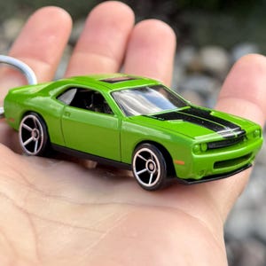 Può includere: Un'auto giocattolo verde con strisce nere sul cofano. Il veicolo in miniatura ha ruote nere e argentate e un tetto apribile. L'auto è un modello Dodge Challenger.