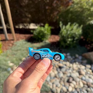 Może przedstawiać: Jasnoniebieski samochodzik z numerem 95 z boku i napisem "Dinoco" na spojlerze. Samoch&oacute;d ma srebrne koła i jest trzymany w dłoni osoby.