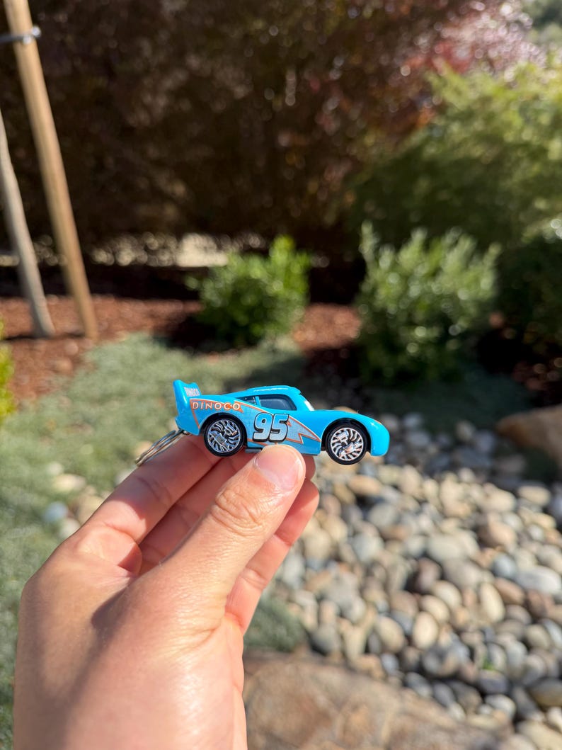 Może przedstawiać: Jasnoniebieski samochodzik z numerem 95 z boku i napisem "Dinoco" na spojlerze. Samoch&oacute;d ma srebrne koła i jest trzymany w dłoni osoby.