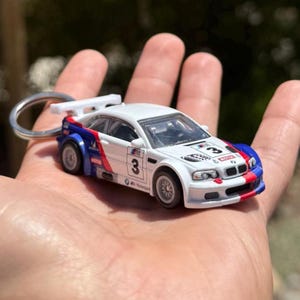 Puede incluir: Un llavero de coche de carreras en miniatura blanco, azul y rojo. El coche tiene el número 3 en el lateral y el capó, con logotipos de patrocinadores. El llavero tiene un anillo plateado y un pequeño alerón.
