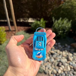 Może przedstawiać: Jasnoniebieski samochodzik z numerem 95 i logo Dinoco. Miniaturowa replika samochodu wyścigowego ma srebrny brelok do kluczy. Samoch&oacute;d jest trzymany w dłoni osoby. Tło jest rozmyte.