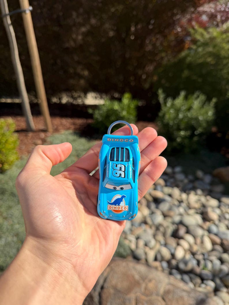 Może przedstawiać: Jasnoniebieski samochodzik z numerem 95 i logo Dinoco. Miniaturowa replika samochodu wyścigowego ma srebrny brelok do kluczy. Samoch&oacute;d jest trzymany w dłoni osoby. Tło jest rozmyte.