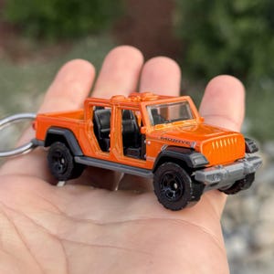 May include: Orange toy Jeep Mojave with black wheels and a silver keychain. The toy car has black seats and a windshield. The word "Mojave" is printed on the side of the car.