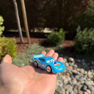 Może przedstawiać: Jasnoniebieski brelok do kluczy w kształcie zabawkowego samochodu z numerem "95" i napisem "Dinoco". Samoch&oacute;d ma uśmiechniętą twarz i jest trzymany w dłoni.