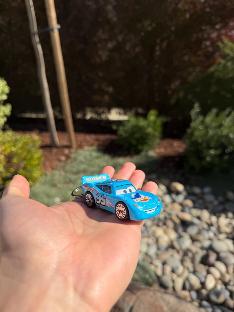Może przedstawiać: Jasnoniebieski brelok do kluczy w kształcie zabawkowego samochodu z numerem "95" i napisem "Dinoco". Samoch&oacute;d ma uśmiechniętą twarz i jest trzymany w dłoni.