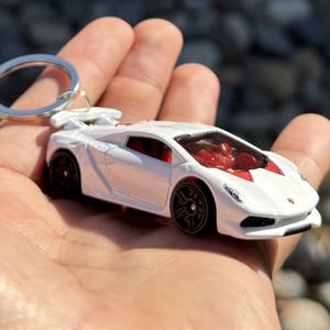 May include: A white toy sports car keychain with a red interior and black wheels. The car has a spoiler and is attached to a silver keyring. The car is held in a person's hand.