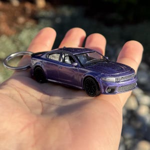 以下が含まれることがあります： 黒い車輪と銀色のキーリングが付いた、ミニチュアの紫色の車のキーホルダー。車には、ヘッドライト、グリル、窓など、詳細なデザインが施されています。車はぼやけた背景に対して手で持たれています。