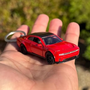 以下が含まれることがあります： 黒い屋根と黒い車輪が付いた赤いおもちゃの車で、銀色のキーホルダーに取り付けられています。 ミニチュアカーはクーペスタイルで、洗練されたデザインです。 車は、ぼやけた緑色の背景に対して、人の手で持たれています。