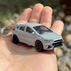 May include: A silver die-cast model car keychain. The miniature car features black wheels and a detailed design, including a front grill and headlights. The keychain ring is silver.