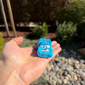 Może przedstawiać: Jasnoniebieski samochodzik z logo Dinoco na masce i bokach. Samoch&oacute;d ma numer 95 na dachu. Samoch&oacute;d trzymany jest w dłoni na rozmytym tle zieleni i kamieni.