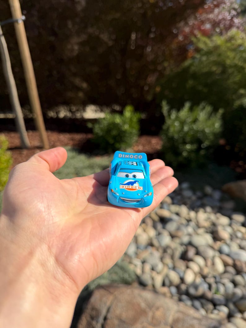 Może przedstawiać: Jasnoniebieski samochodzik z logo Dinoco na masce i bokach. Samoch&oacute;d ma numer 95 na dachu. Samoch&oacute;d trzymany jest w dłoni na rozmytym tle zieleni i kamieni.