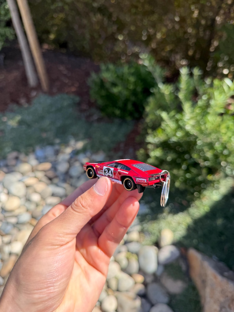 May include: A red toy car with black and white accents, featuring the number 34 on the side. The car has a keychain attached. The background is blurred, with green foliage and rocks.