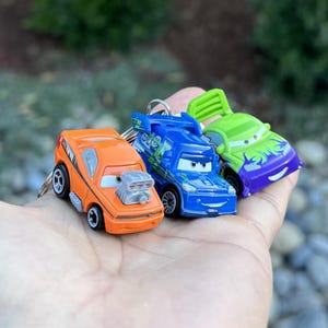 Puede incluir: Tres llaveros de coches de juguete en naranja, azul y verde. El coche naranja tiene un motor plateado y ruedas negras. El coche azul tiene un alerón y detalles verdes. El coche verde tiene detalles morados. Los tres tienen caras sonrientes.