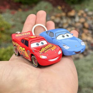 May include: Two toy cars from the movie Cars. One is red with the number 95 and the name "Rusteze" on it. The other is a light blue car. Both have cartoon eyes and are keychains.
