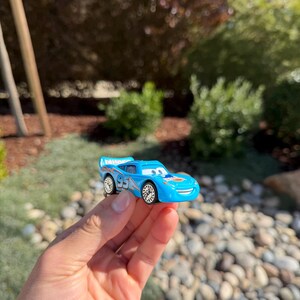 Może przedstawiać: Jasnoniebieski zabawkowy samoch&oacute;d wyścigowy z numerem "95" z boku. Samoch&oacute;d ma uśmiechniętą twarz i spojler. Koła są srebrne. Samoch&oacute;d jest trzymany w dłoni na tle zieleni i skał.